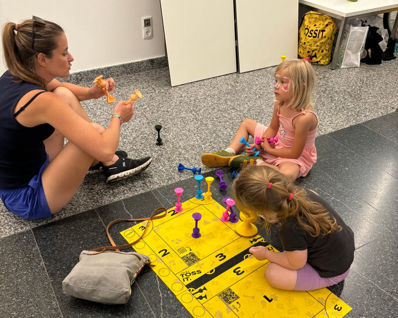 A woman and two children sitting on the floor playing on a yellow TOSSIT mat, coloured darts across the numbered squares
